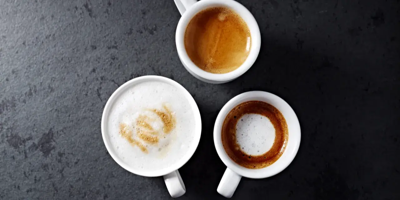 Trzy białe filiżanki z kawą: espresso, latte macchiato i kawą z pianką na ciemnym tle.