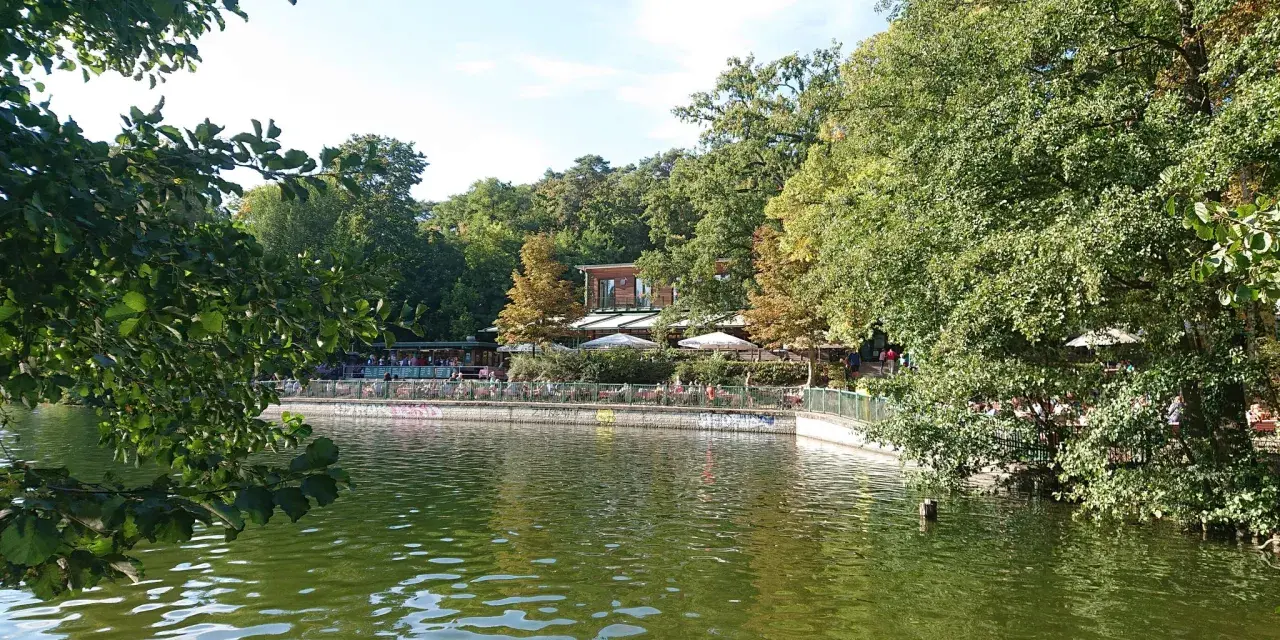 Grüne Bäume säumen den Schlachtensee Berlin, wo Menschen auf der Terrasse eines Restaurants sitzen.