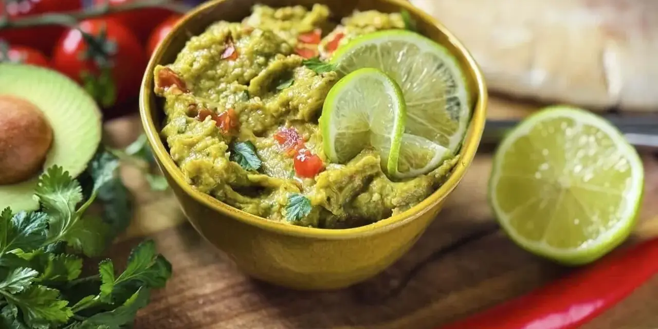 Frisches Guacamole-Rezept in einer gelben Schüssel, garniert mit Limettenscheiben. Daneben liegen Avocado, Koriander, Tomaten und Chili.