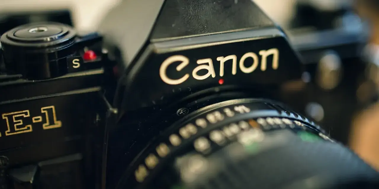 Zbliżenie na czarną lustrzankę Canon AE-1. Widoczny napis "Canon" i fragment obiektywu. Idealny do połączenia z aparatem.