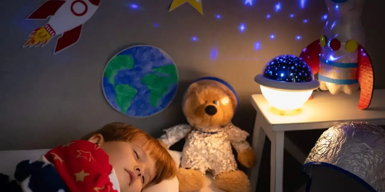 Śpiące dziecko pod kocem w gwiazdki, obok pluszowy miś w stroju astronauty. Na ścianie rakieta i Ziemia, a na suficie projektor dla dzieci na sufit wyświetla gwiazdy.