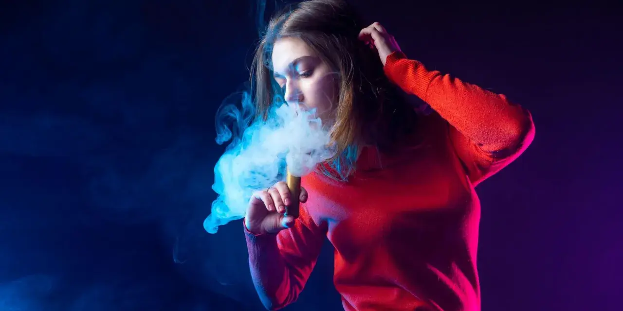 Joven exhala humo de un vaper, rodeada de luces azules y moradas.