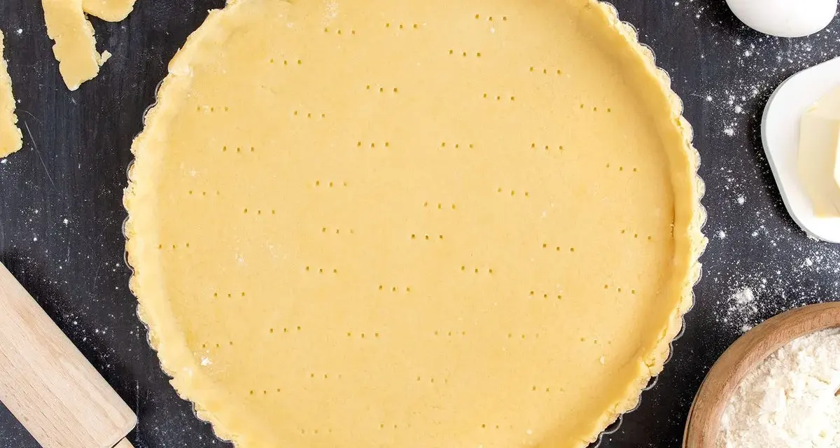 Ein vorbereiteter Mürbeteigboden in einer Tarteform, bereit für das 1 2 3 Teig Rezept. Zutaten wie Eier, Butter und Mehl sind daneben zu sehen.