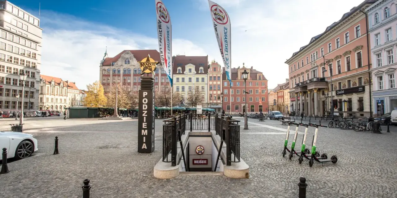 Wejście do podziemi z napisem "Movie Gate Wrocław" na placu w otoczeniu zabytkowych kamienic.