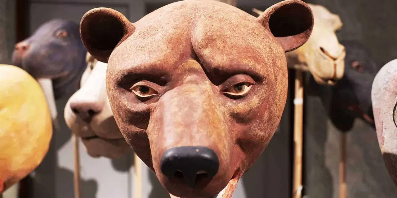 En brun björnmans ansikte, gjord av papier mache lim, med andra djurmasker i bakgrunden.
