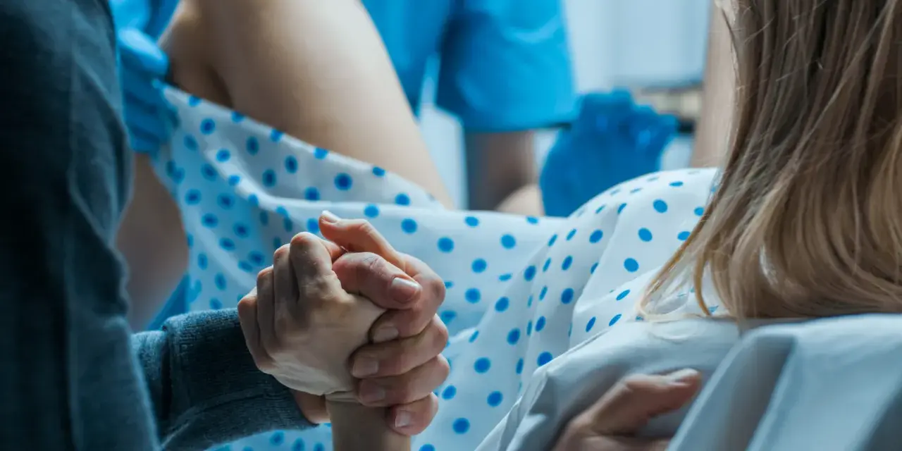 Wsparcie bliskiej osoby podczas porodu. Dłoń partnera trzyma dłoń rodzącej, która ma już widoczny czop śluzowy.