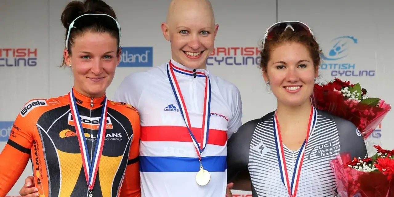Joanna Rowsell, a bald cyclist in a white and red jersey, smiles on the podium with two other medalists.