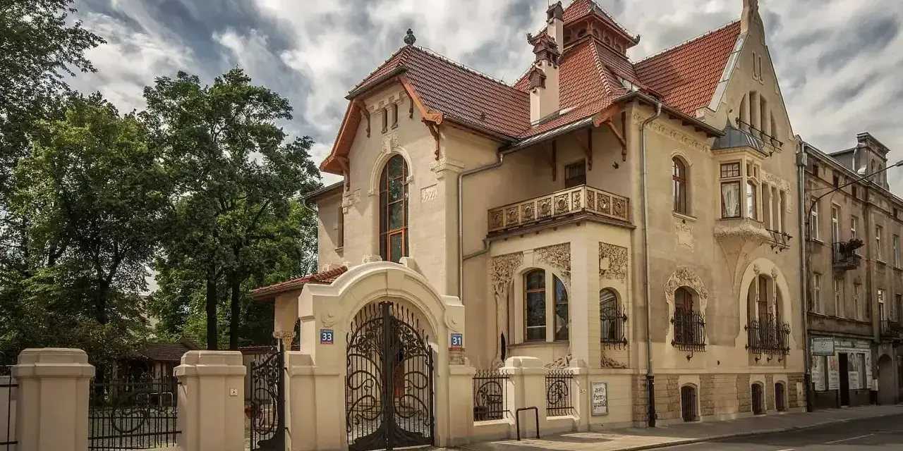 Elegancka willa w stylu art nouveau z bogatymi zdobieniami, kutą bramą i ozdobnym balkonem.