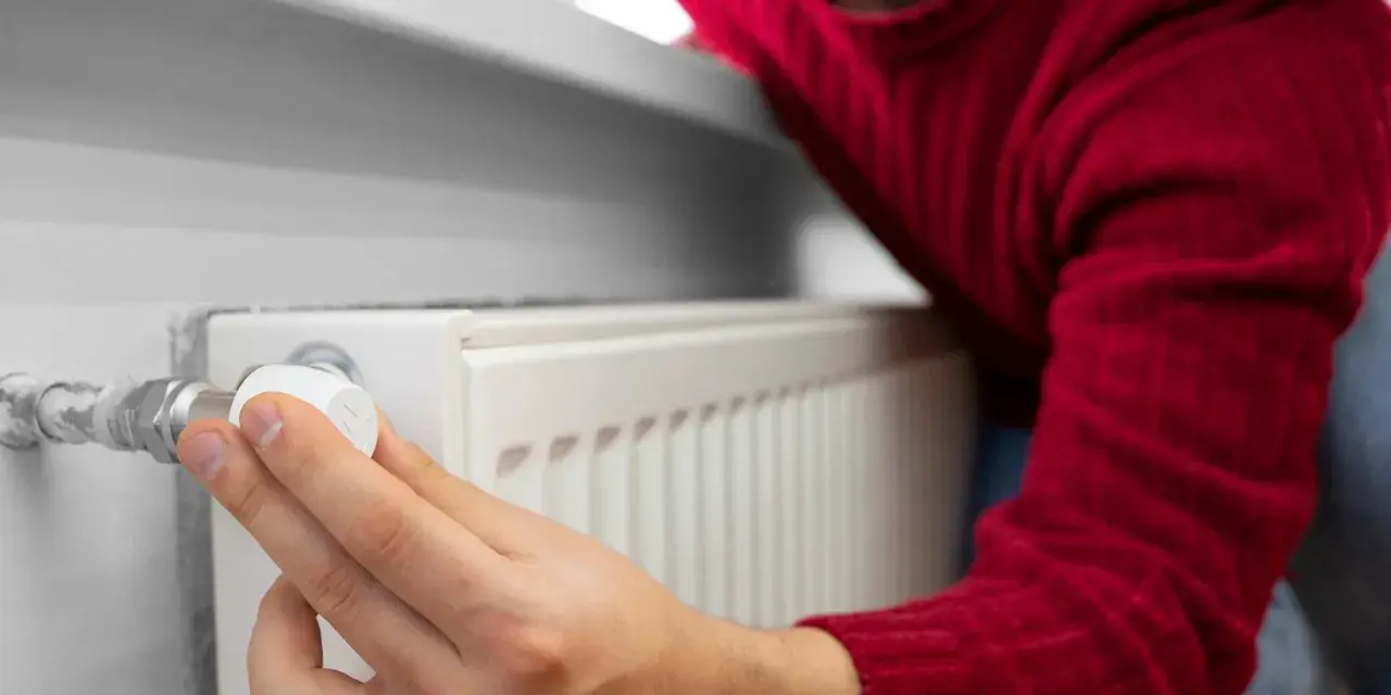 Mężczyzna w czerwonym swetrze reguluje termostat grzejnika, oszczędzając energię. To jego sposób na wykorzystanie bonu ciepłowniczy.