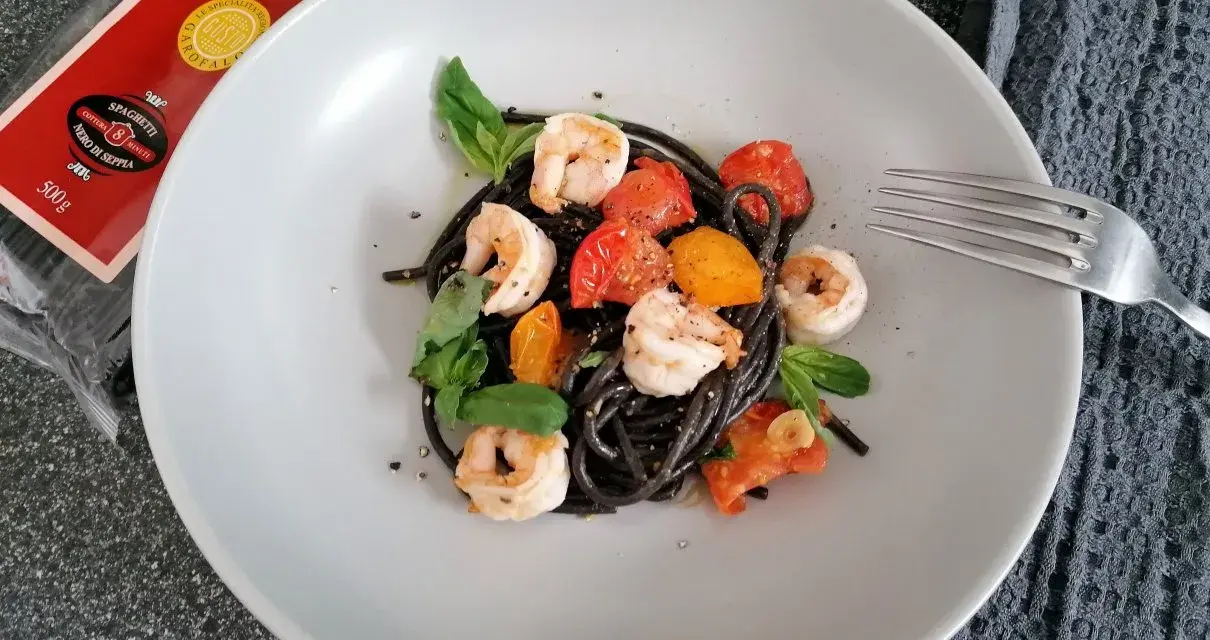 Czarny makaron z krewetkami, pomidorkami i bazylią na talerzu. Obok widoczna paczka spaghetti nero di seppia.