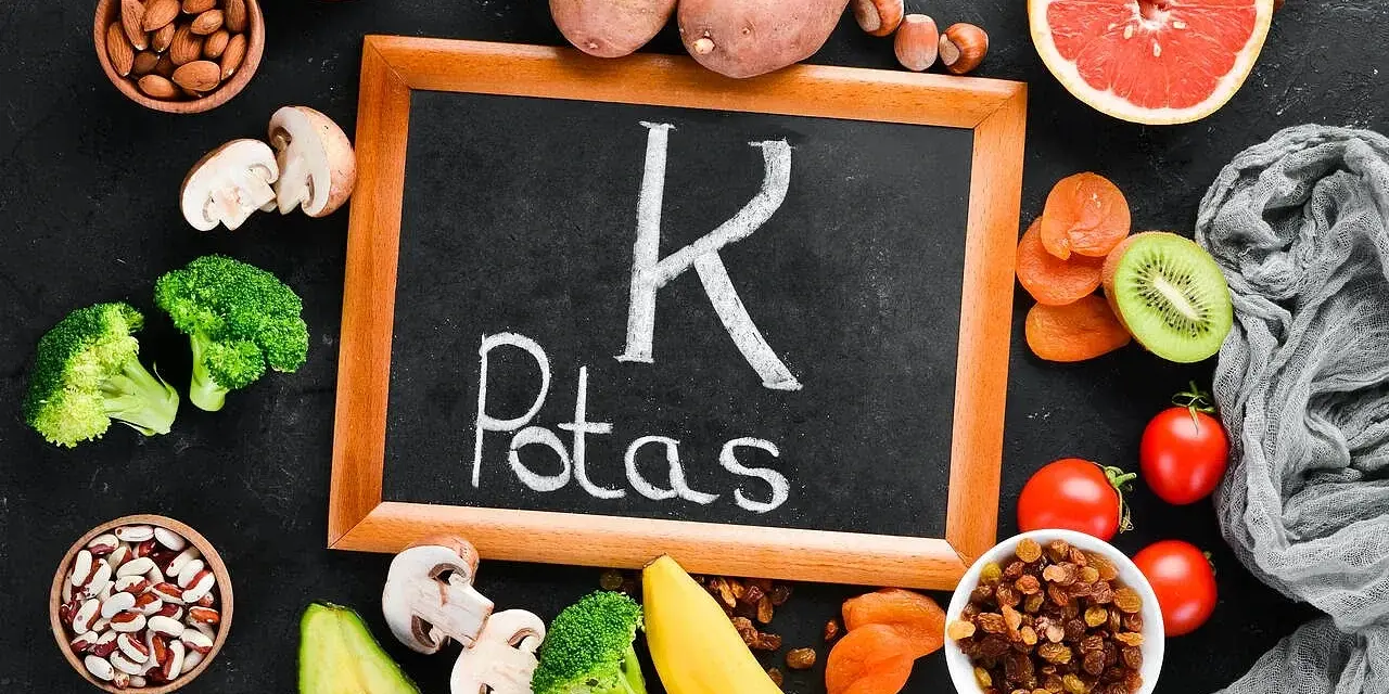 Tablica z napisem "K Potas" otoczona owocami i warzywami bogatymi w potas, jak banany, awokado, pomidory, ziemniaki, brokuły, rodzynki, morele, kiwi, migdały, pestki dyni. Pomaga zapobiegać hipokaliemii.