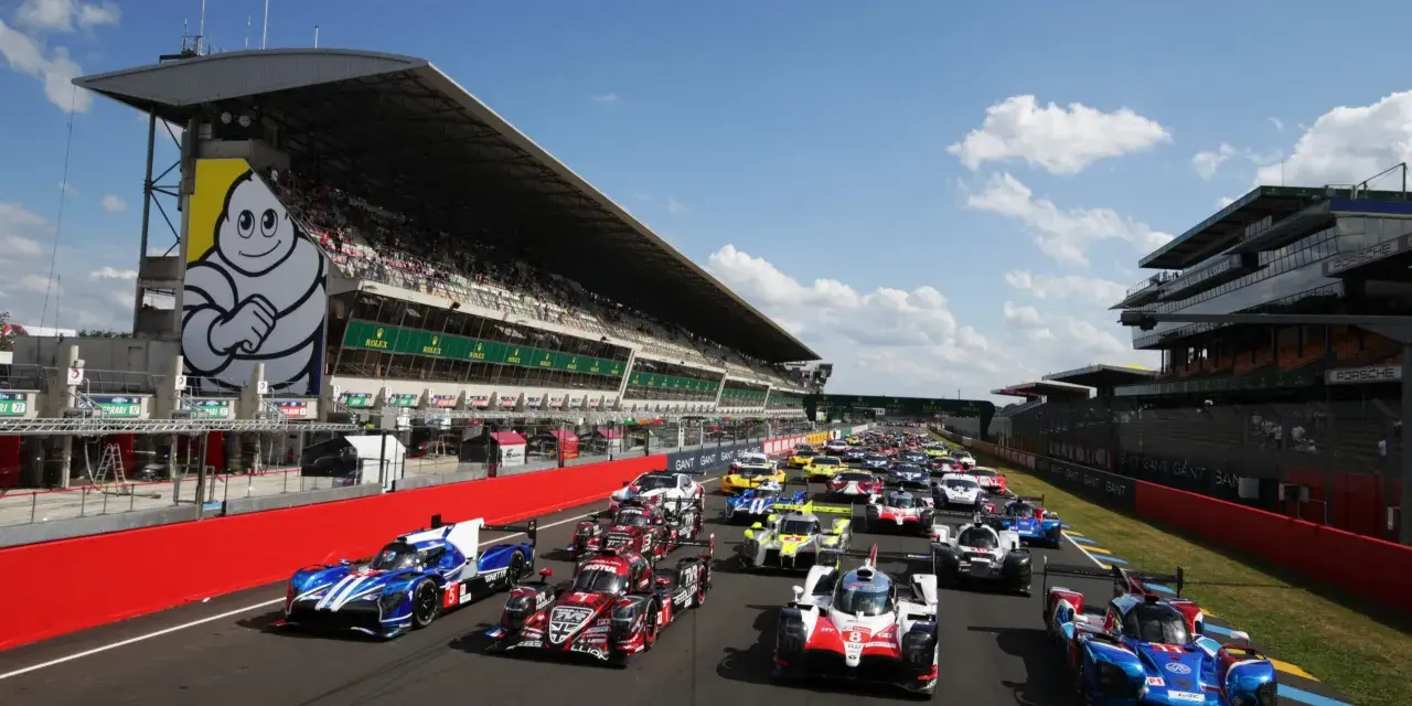 Una fila de coches de carreras listos para comenzar las 24 horas de Le Mans. El icónico muñeco de Michelin preside la tribuna.