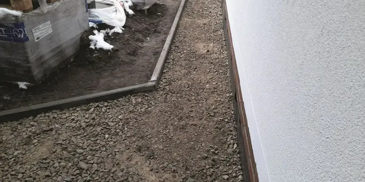 Wykonano opaskę wokół domu z kruszywa, obok ułożone palety z materiałami budowlanymi.