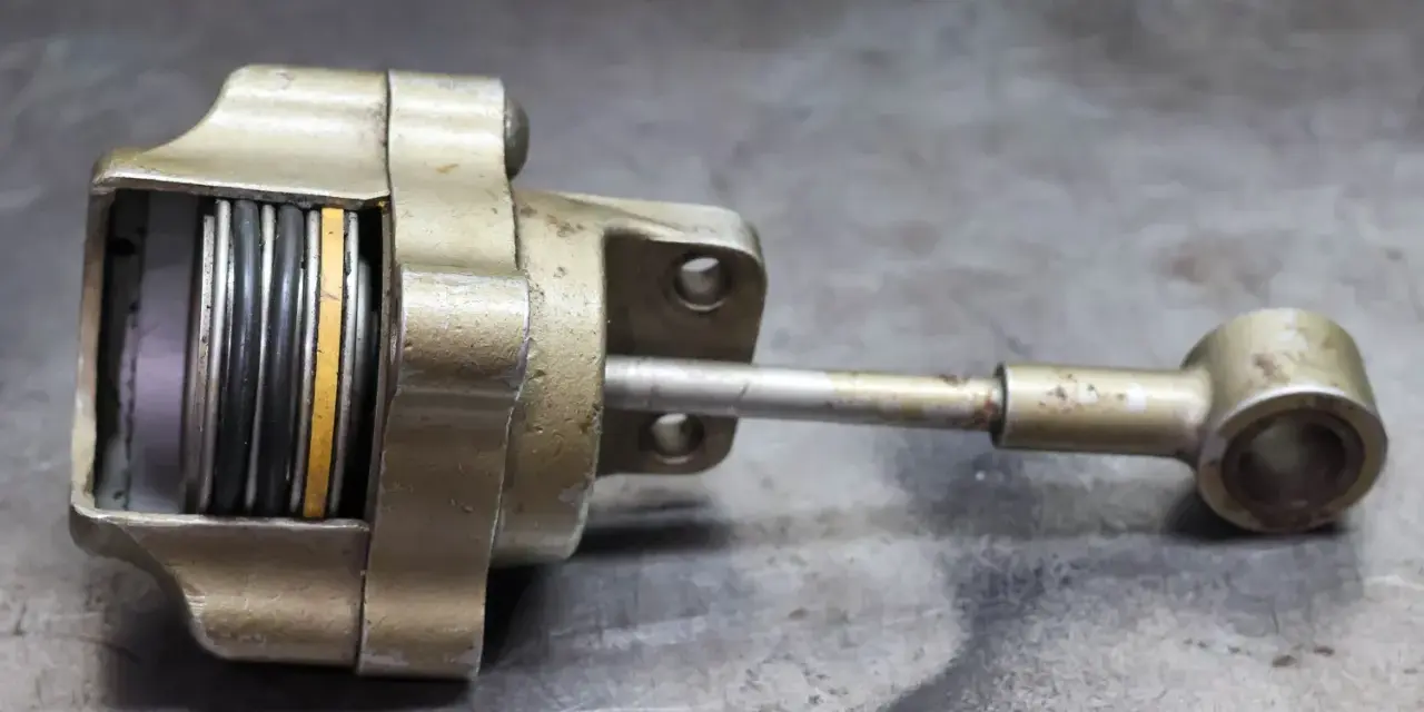 Metalowy element z tłoczyskiem i sprężynami, przypominający zawór wastegate. Objawy uszkodzenia mogą być widoczne na jego powierzchni.