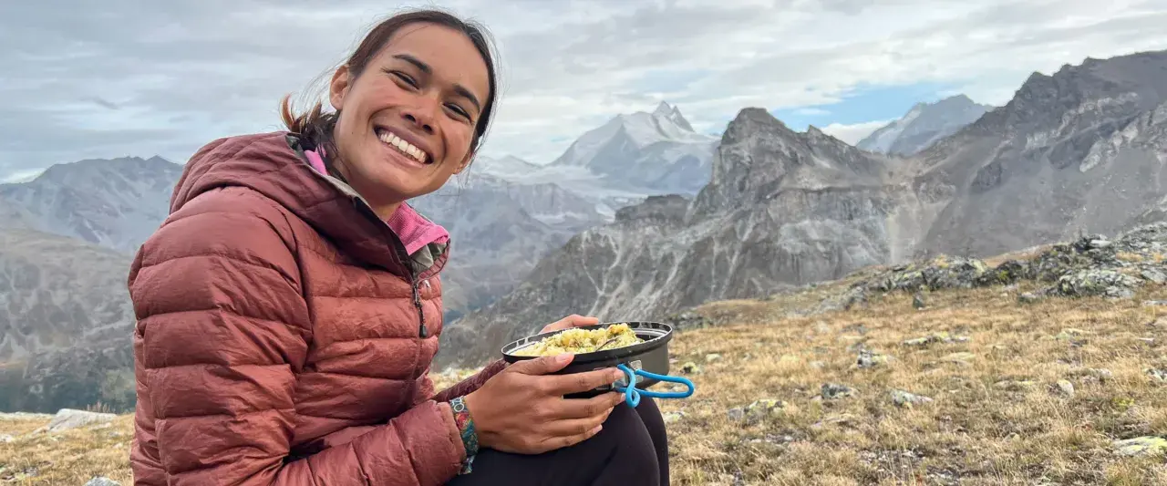 Sourire radieux d'une randonneuse savourant son repas en pleine montagne, avec des sommets enneigés en arrière-plan. Un moment de pur bonheur pour ce repas randonnée.