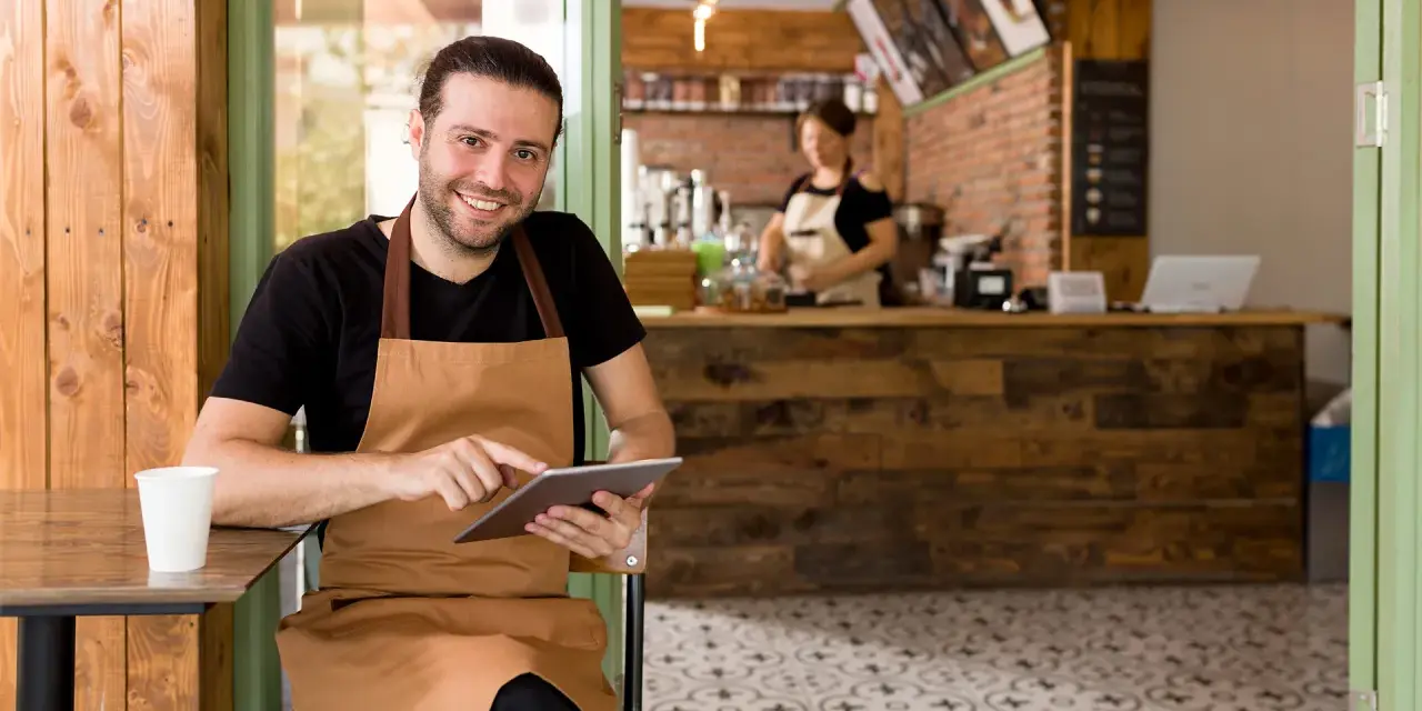 Hombre sonriente con delantal usa tablet en su cafetería, gestionando pedidos con un sistema erp restaurantes.