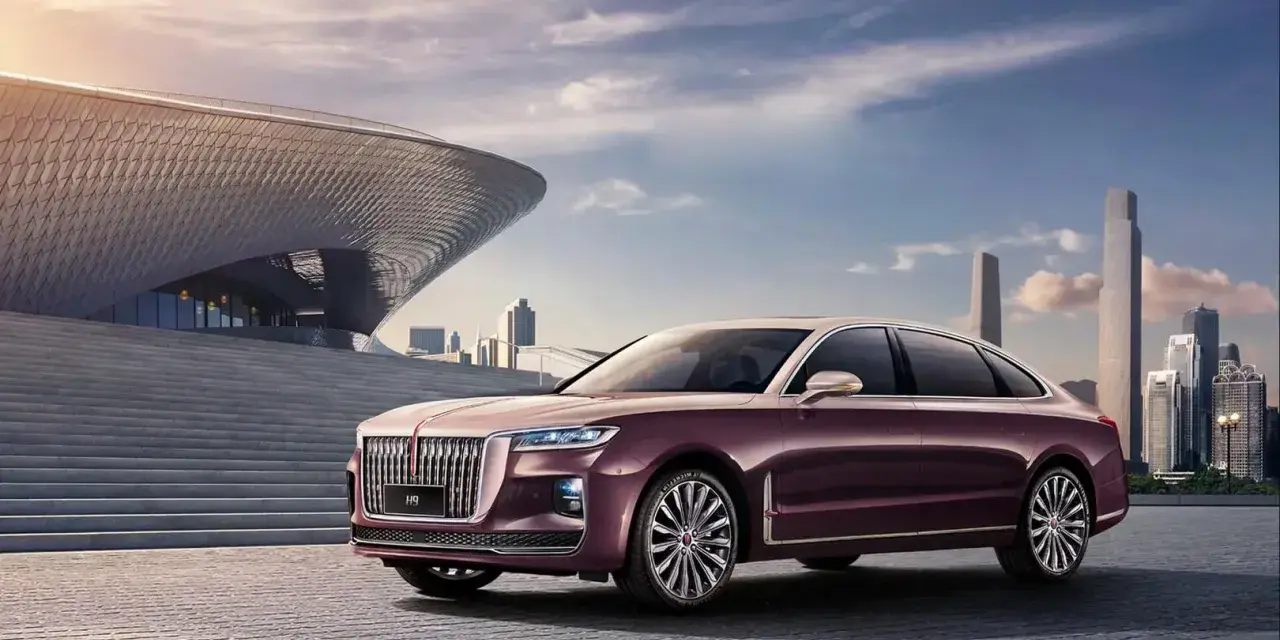 Elegancki, bordowy Hongqi H9 z chromowanymi detalami stoi przed nowoczesnym budynkiem o falistej fasadzie, w tle miejski krajobraz.