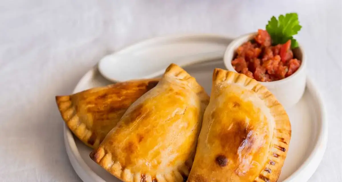 Tres empanadillas al horno doradas y crujientes, servidas en un plato blanco con un pequeño cuenco de salsa de tomate y una hoja de perejil.