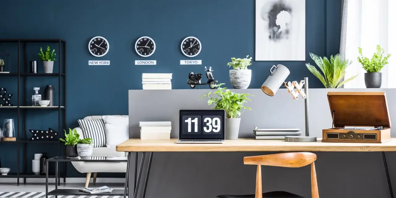 Biuro z zegarami pokazującymi czas w Nowym Jorku, Londynie i Tokio na ścianie w kolorze navy. Na biurku laptop, lampa i gramofon.