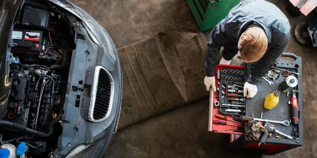 Meccanico con guanti bianchi esamina un motore sbiellato, circondato da attrezzi e un'auto con il cofano aperto.