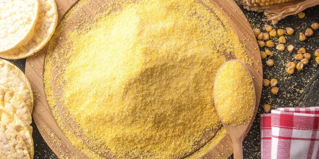 Stos mąki kukurydzianej na desce, obok łyżka z mąką, kukurydza i wafle.