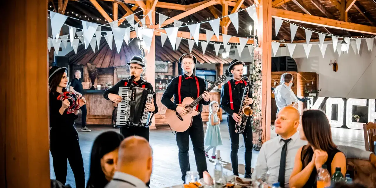 Zespół grający na weselu, z akordeonem, gitarą i saksofonem. W tle widać gości i dekoracje. Idealni do oprawy muzycznej imprez, znają teksty piosenek biesiadnych i weselnych.