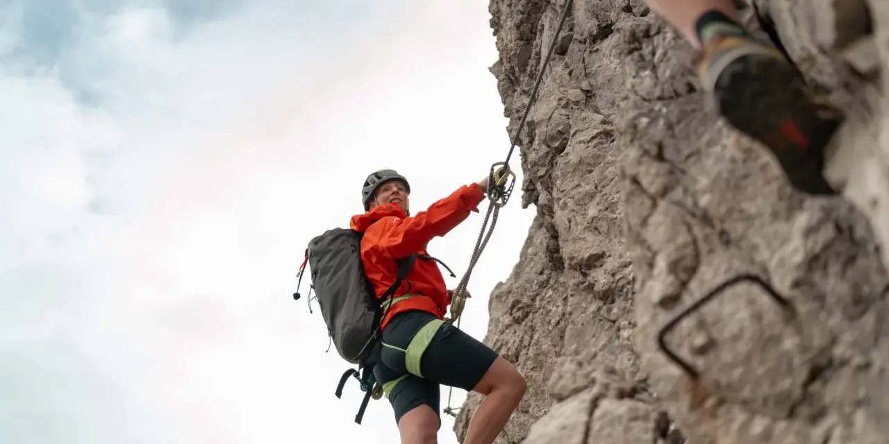 Kletterin im orangefarbenen Anzug sichert sich am Drahtseil des Alpspitz Klettersteigs. Ein spannendes Abenteuer in den Bergen.