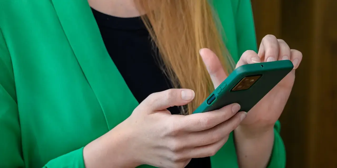 Kobieta w zielonej marynarce trzyma telefon. Szuka informacji, gdzie jest schowek w telefonie, aby ukryć ważne dane.