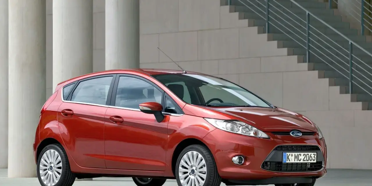 Czerwony Ford Fiesta MK6 zaparkowany przed nowoczesnym budynkiem z betonowymi kolumnami i schodami.