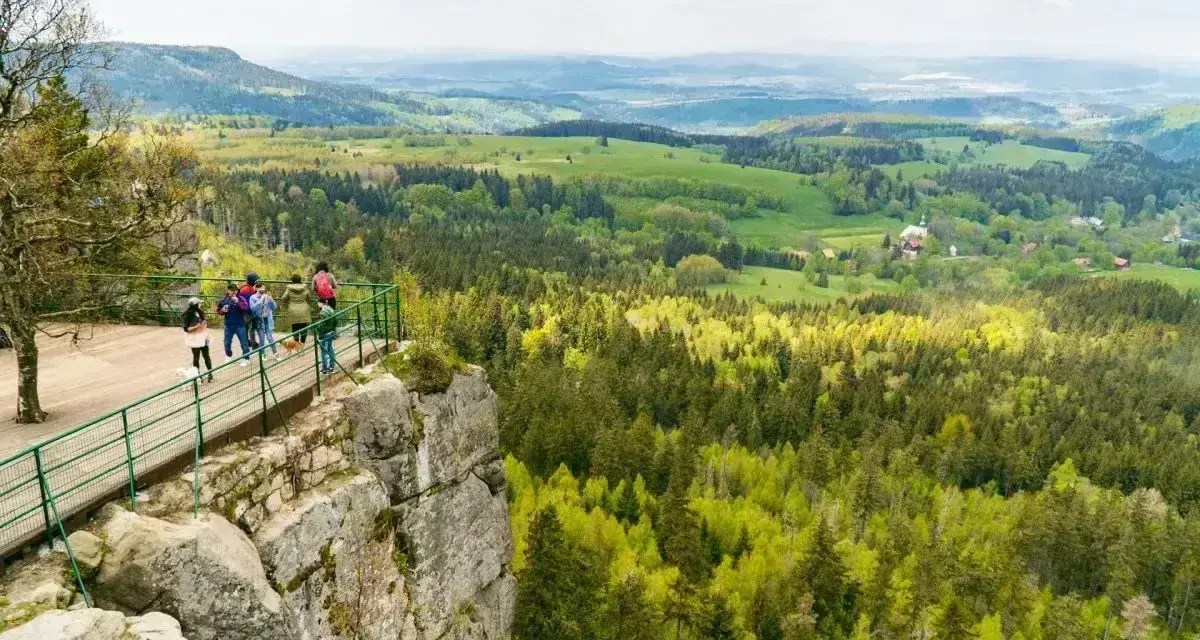 Grupa turystów podziwia rozległą panoramę Kotliny Kłodzkiej z platformy widokowej na skalnym urwisku.