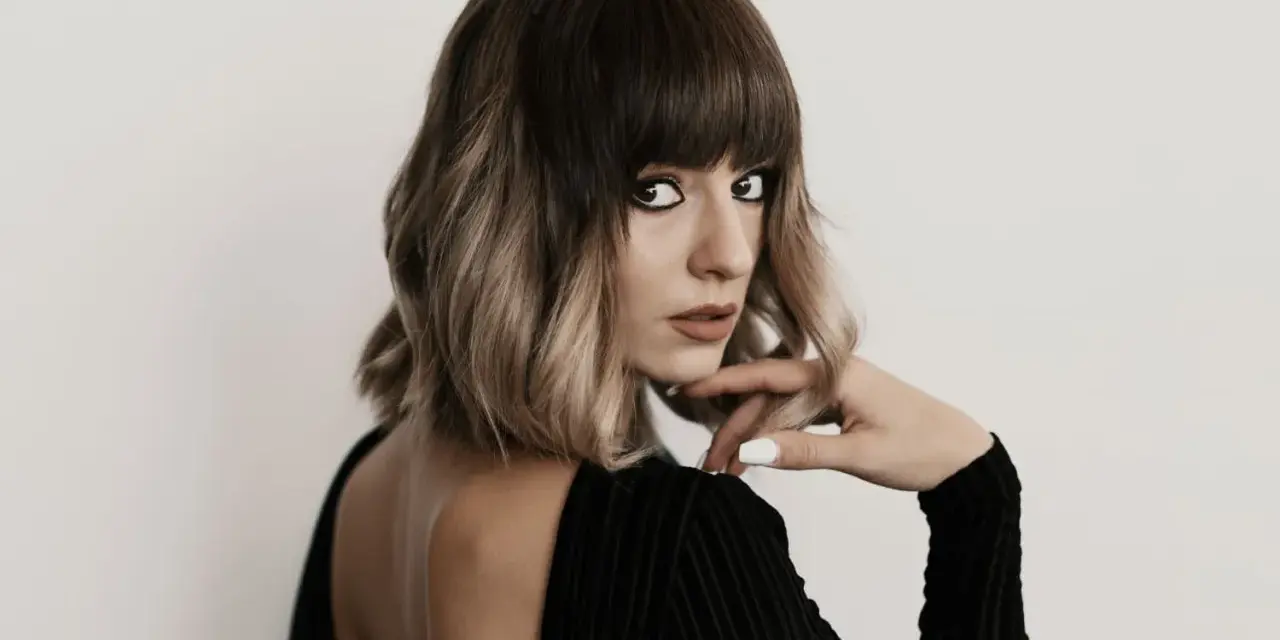 Kobieta z modnym, **blunt bob** w odcieniach brązu i blondu, z grzywką, w czarnej sukience z odkrytymi plecami.