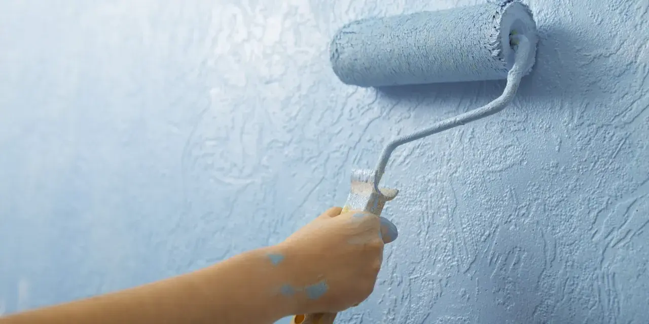 Mano aplica imprimación azul con rodillo a pared texturizada. La imprimación es el primer paso para un acabado perfecto.