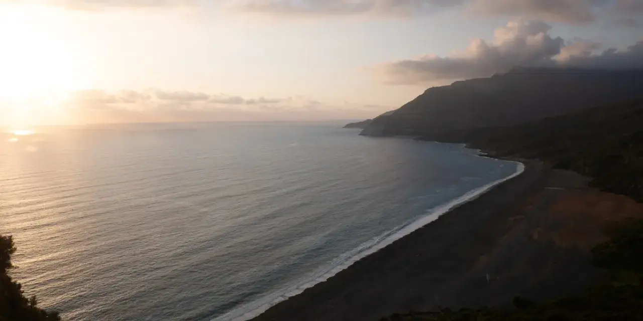 Vue aérienne d'une plage de sable noir bordée par une mer calme et des montagnes embrumées. Ces photos de plage capturent la sérénité d'un paysage côtier au coucher du soleil.