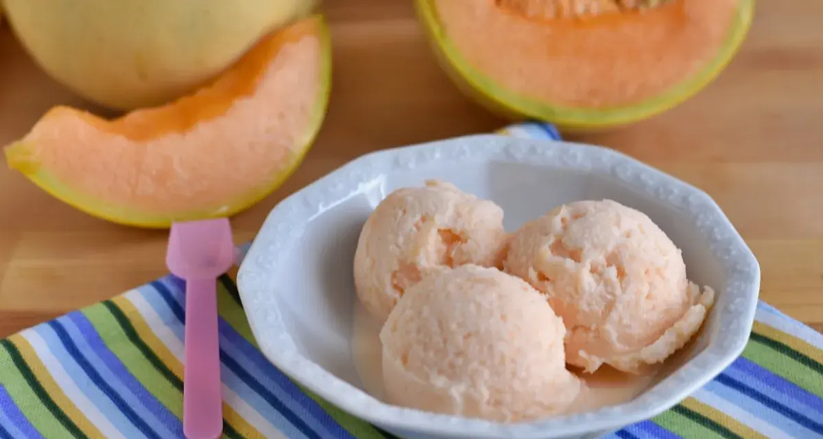 Tre palline di cremoso gelato al melone in una ciotola bianca, con fette di melone fresco sullo sfondo.