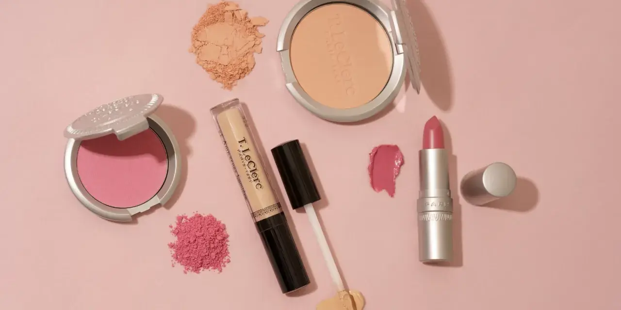 Un assortiment de maquillage rose et doré : blush, poudre, fond de teint, rouge à lèvres et fard à paupières sur fond rose.