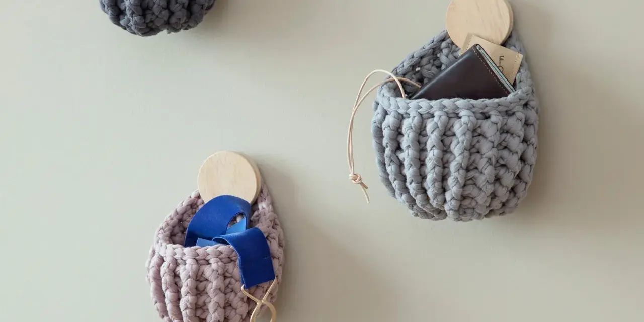 Three crocheted wall pockets in grey and pink, one holding a wallet, another blue ribbon, on a light wall.