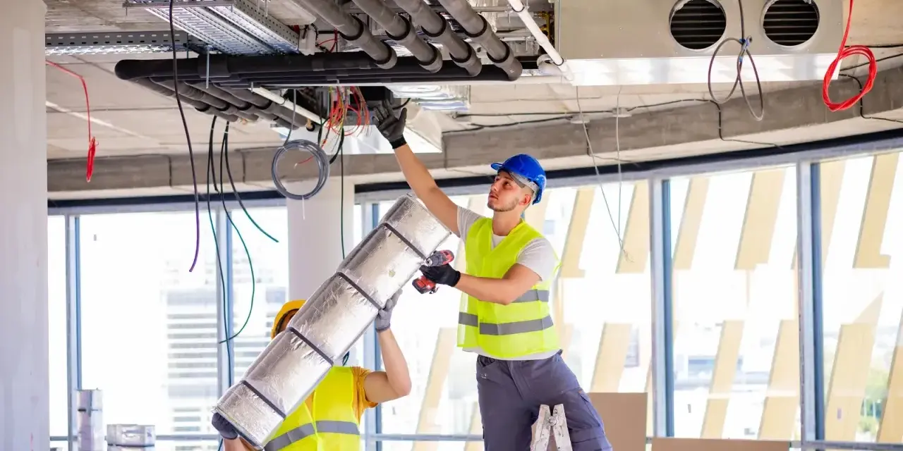 Dwóch pracowników montuje systemy HVAC, instalując izolację na rurach. To kluczowy element zapewniający komfort cieplny.