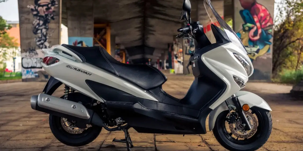 Biały skuter Suzuki Burgman 125, z czarnym siedzeniem, zaparkowany pod mostem z kolorowymi graffiti. Idealny do miasta, sprawdź suzuki burgman 125 dane techniczne.