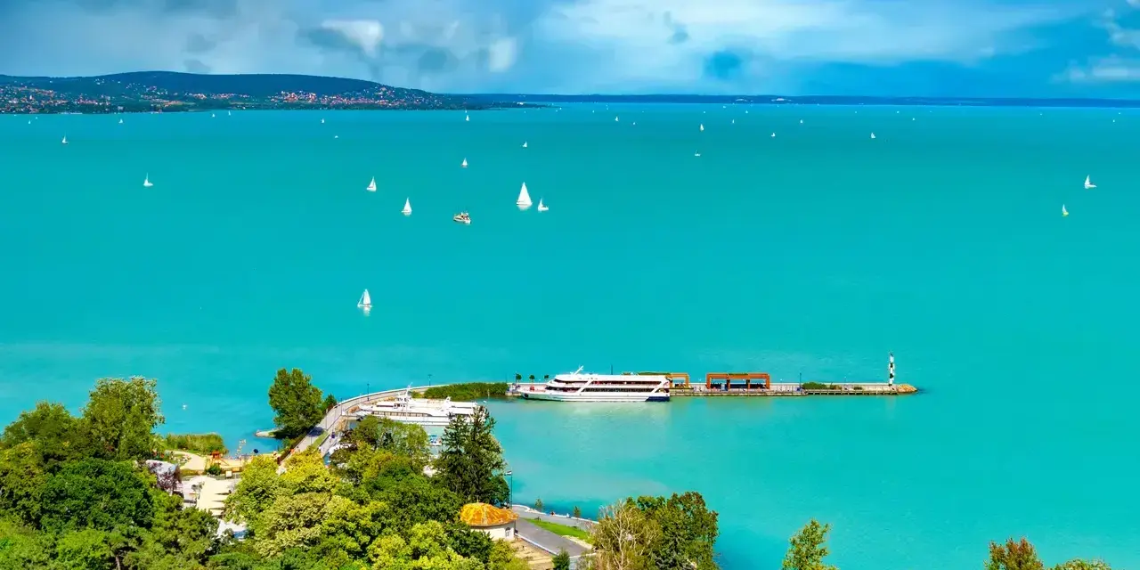 Turkusowe wody jeziora Balaton, pełne żaglówek i statków, z zielonymi wzgórzami w tle.