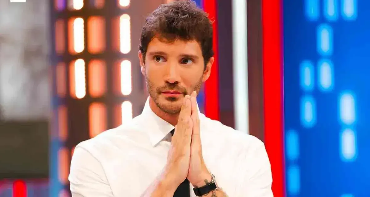 Santiago de Martino, con le mani giunte in preghiera, guarda intensamente verso l'obiettivo, con uno sfondo di luci colorate.