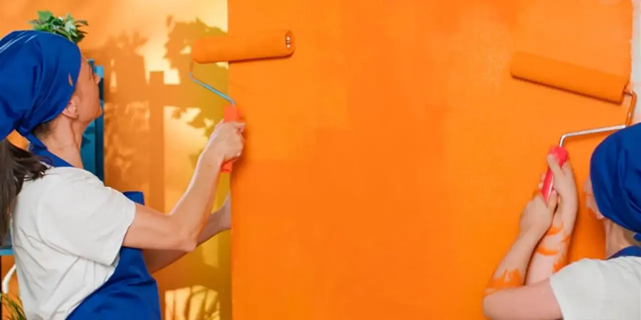 Dos personas pintan una pared de naranja brillante con rodillos. La pintura mural exterior cobra vida con vibrantes tonos anaranjados.