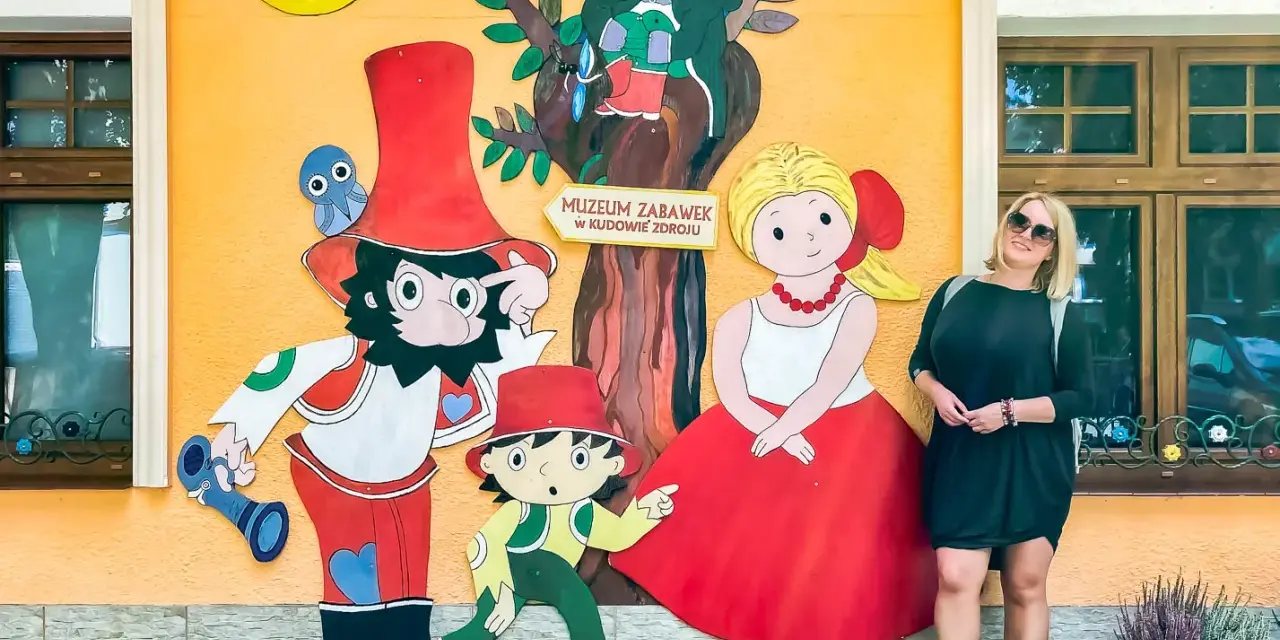 Kobieta pozuje przy kolorowej ścianie Muzeum Zabawek w Kudowie-Zdroju, Dolny Śląsk. Wesołe postacie z bajek towarzyszą jej w radosnej scenerii.