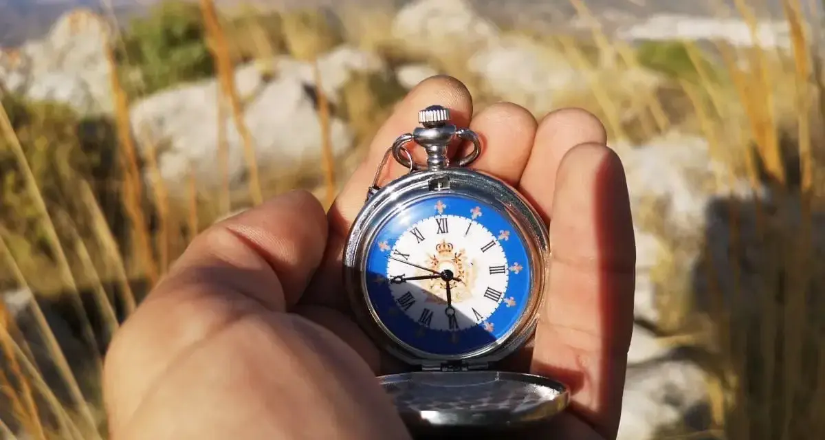 Mano sostiene un reloj de bolsillo antiguo con esfera azul y números romanos, útil para orientarse con reloj de agujas en un paisaje montañoso.