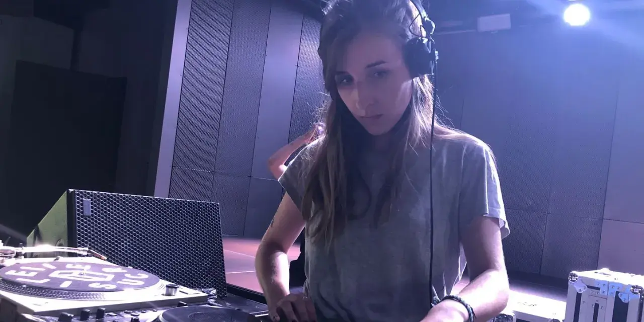 Une dj fille concentrée aux platines, casque sur les oreilles, dans une ambiance tamisée avec des lumières bleues.