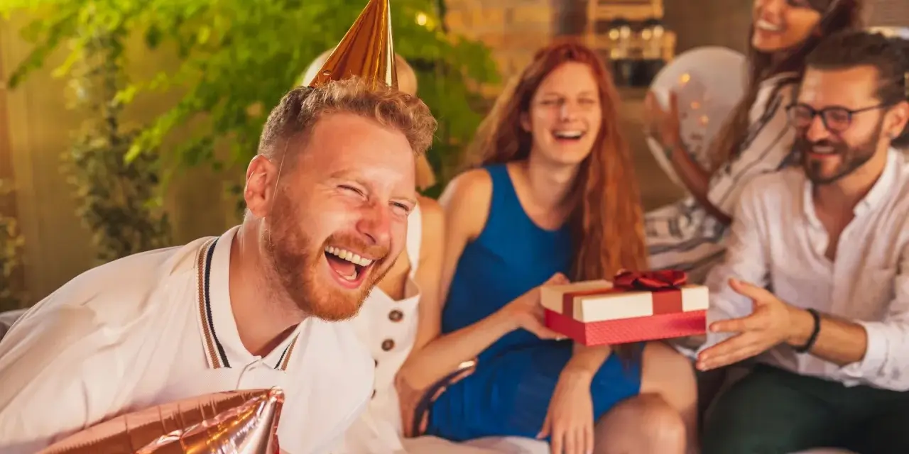 Radosna ekipa świętuje urodziny. Wszyscy się śmieją, a solenizant w złotym czepku trzyma balon. Czas na sto lat śmieszne życzenia!