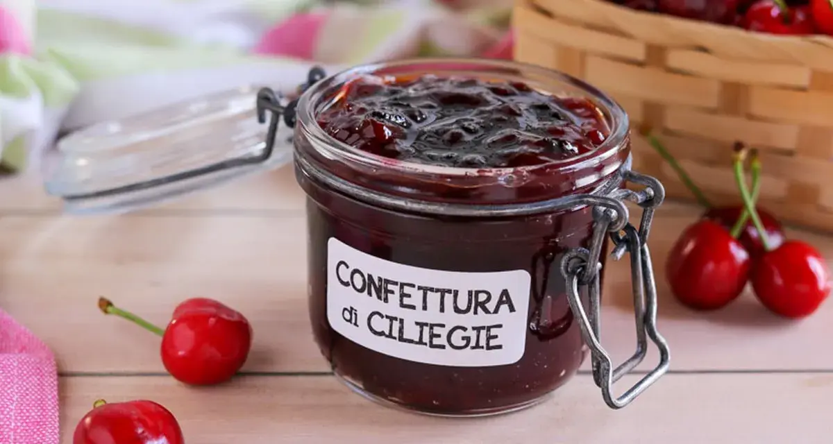 Un vasetto di marmellata di ciliegie fatta in casa, con ciliegie fresche e un cestino sullo sfondo.