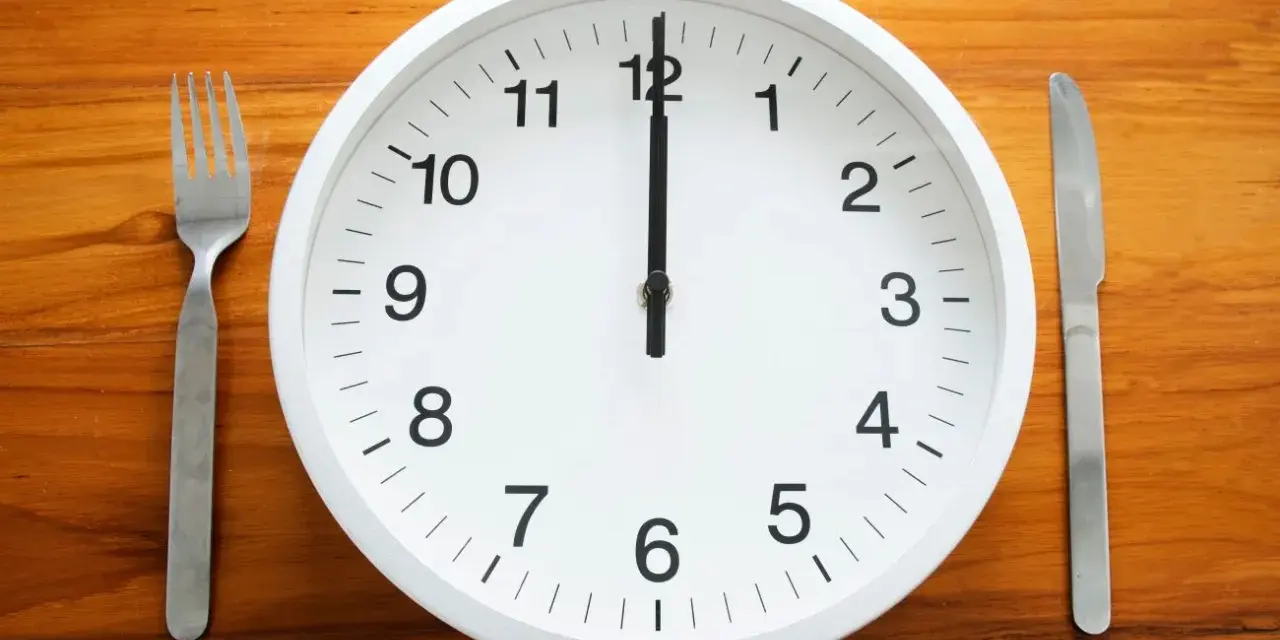 Uhr zeigt 12 Uhr. Gabel und Messer liegen bereit. Zeit für die Mittagspause während der Arbeitszeit.