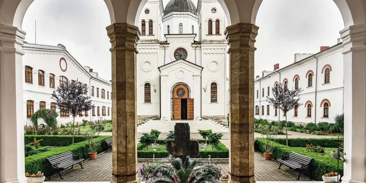 A courtyard with a cross and potted plants, framed by arches. The white church in the background is a place where monks like Father John and Brother Michael find peace.