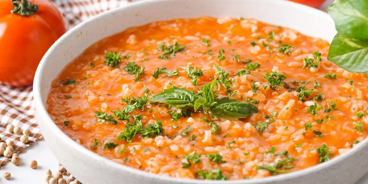 Eine cremige Tomatensuppe mit Reis, garniert mit frischer Petersilie und Basilikum. Daneben liegen frische Tomaten und Gewürzkörner.