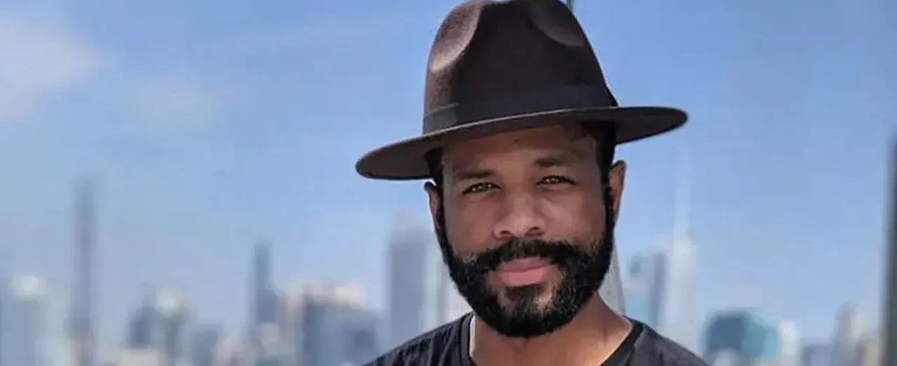 Hombre con sombrero y barba espesa, sonriendo ante un fondo de edificios y cielo azul.